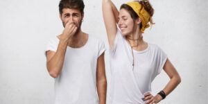 Problems with body odor. Disgusted male pinching his nose feeling bad smell or stink coming out from attractive smiling girl, who is raising her arm, showing wet t-shirt because of armpit sweat